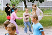 Динамічна вулична сцена з кількома дітьми, які граються. У центрі дівчинка в рожевій кофті з довгими рукавами та фіолетових штанях тягнеться до іншої дівчинки у світло-рожевій кофті та синіх джинсах, яка посміхається і має червоні сонцезахисні окуляри на голові. Дівчинка в джинсах перебуває в русі. Червона повітряна кулька лежить на землі ліворуч, а білу повітряну кульку тримає дівчинка праворуч, яка частково закрита. На задньому плані видно інших дітей, зокрема хлопчиків у чорному худі та синій футболці. Дія відбувається на брукованій ділянці поруч із трав'яним газоном, деревом та багатоповерховою будівлею на задньому плані.