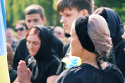  Жінка з сивим волоссям, зібраним у зачіску, у чорній хустці на голові та чорному одязі стоїть, повернувшись у профіль. За нею стоять інші люди, дехто з яких також у чорному одязі, а одна людина складає руки у молитві. Частина синьо-жовтого прапора видно ліворуч.