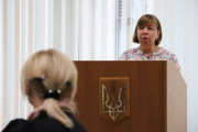 Директор Департаменту культури, туризму, молоді та спорту Наталія Петрович доповідає про проєкт рішення, винесений на засідання виконавчого комітету за трибуною. У центрі кадру знаходиться дерев'яна трибуна світло-коричневого кольору, на передній панелі якої розміщено металевий герб України – тризуб. Позаду жінки білі шпалери, що закривають панрамне вікно. 