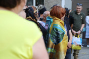  На фотографії зображена група людей, що стоять на вулиці. В центрі знімка знаходиться жінка з рудим волоссям, яка обгорнута жовто-синім прапором. На прапорі зображено герб з емблемою, що включає сокола, що летить над щитом.  Ліворуч від неї стоїть жінка в чорній хустці, праворуч – чоловік у формі з нечітким обличчям та чоловік у синій футболці, на якій видно чиєсь зображення. На передньому плані видно плече та частину спини жінки в салатовій футболці, що стоїть спиною до камери.