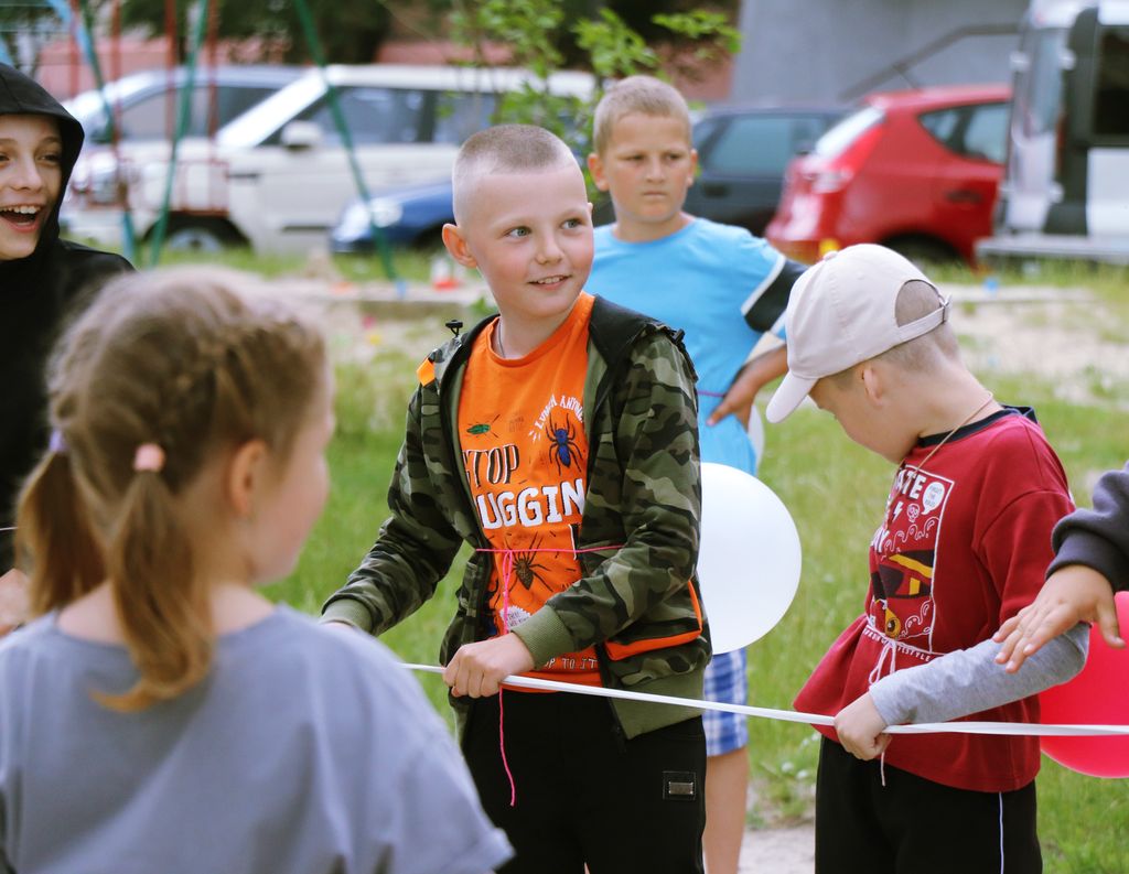 Крупний план кількох дітей, які беруть участь у грі на свіжому повітрі. На передньому плані хлопчик з короткою стрижкою посміхається, дивлячись ліворуч. На ньому помаранчева футболка з малюнком павука під камуфляжною курткою. Праворуч від нього інший хлопчик у білій бейсболці дивиться вниз. Між ними видно білу повітряну кульку. Ліворуч частково видно дівчинку з каштановим волоссям, яка стоїть спиною. На задньому плані інший хлопчик у синій футболці дивиться праворуч, а також припарковані різні автомобілі.