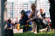  На зображенні показано групу людей на вулиці, які схилились або стоять на колінах. На передньому плані жінка в хустці та темному одязі,  Українською: На зображенні показано групу людей на вулиці, які схилились або стоять на колінах. На передньому плані жінка в хустці та темному одязі, щось витирає біля обличчя і тримає в руках букет помаранчевих лілій. Поруч з нею чоловік на колінах, одягнений у смугасту сорочку та темні шорти, який тримається за груди. За ними видніється ще один чоловік, що також прихилився. На задньому плані видно багатоквартирні будинки та групу людей. Загальна атмосфера зображення виглядає скорботною.