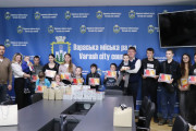 На фотографії зображена група дорослих та дітей, які стоять в приміщенні на фоні синього банера з написом українською та англійською мовами: "Вараська міська рада" та "Varash city council". У верхній частині банера розміщено герб міста Вараш. Зліва на банері також видно частину державного прапора України – жовту та блакитну смуги.  У центрі переднього плану на столі лежать стопки світлих коробок та кілька паперових пакетів білого кольору.  У першому ряду стоять діти різного віку, тримаючи в руках невеликі планшети в чохлах та, ймовірно, солодкі подарунки в прозорих упаковках. Діти посміхаються.  У другому ряду стоять дорослі. З лівого краю знаходяться заступник міського голови Роман Хондока та начальник служби у справах дітей Ольга Самохіна, яка тримає на руках дитину. Ближче до центру, правіше від них, розташований міський голова Олександр Мензул, який також тримає на руках маленьку дитину. Інші дорослі стоять поруч та позаду, також посміхаючись у напрямку камери.  Освітлення в приміщенні яскраве, штучне. Загальна атмосфера на фотографії позитивна та радісна, що відображає подію вручення подарунків дітям.